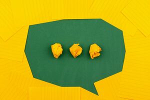 Grüne Sprechblase mit drei zerknüllten gelben Papierkugeln darin, umgeben von gelben Papierblättern.